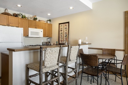 A small kitchen with light wooden cabinets, a white microwave, stove, and fridge. A breakfast bar with three upholstered stools and a round table with four chairs are in the foreground.