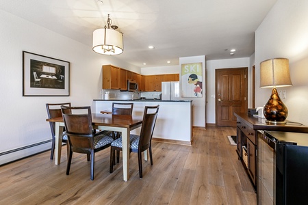 A modern kitchen and dining area with wooden cabinets, a dining table with four chairs, a poster on the wall, and a lamp on a side table. The room has light-colored walls and wood flooring.