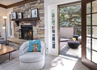 A cozy living room with a stone fireplace, a gray armchair featuring a deer pillow, and a view of an outdoor patio with a fire pit through open glass doors.