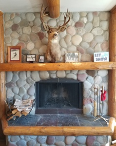 Stone fireplace with a mounted deer head above the mantel. The mantel holds various framed pictures, a "Let it Snow" sign, and other small decorations. Firewood and fireplace tools are nearby.