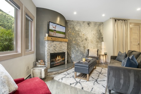 Modern living room with a fireplace, mounted TV, and comfortable seating. Large windows provide a view of greenery, and a mix of textured walls adds to the contemporary decor.