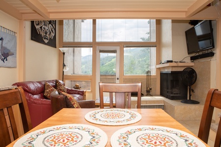 A cozy living area with wooden furniture, a red leather sofa, a fireplace, and a wall-mounted TV. Large windows and a sliding door offer a view of a green mountain landscape.