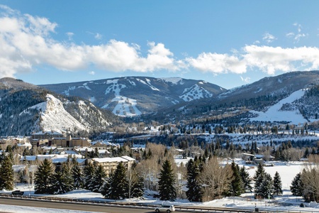 A snow-covered mountainous landscape with a ski resort area, residential buildings, and pine trees in the foreground under a clear blue sky.