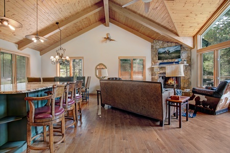 A spacious living room with wooden vaulted ceilings, a stone fireplace, and large windows. The room features a bar area with stools, a sofa, an armchair, and a chandelier.