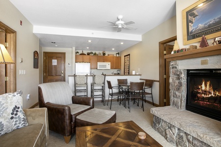 A cozy living room with a stone fireplace, a leather armchair, and a sofa. A dining table with four chairs is near the open kitchen, which has wooden cabinets, a microwave, and a refrigerator.