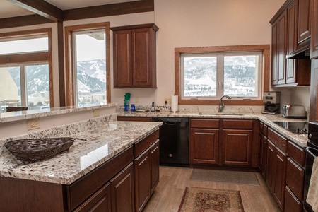 A kitchen with dark wood cabinets, granite countertops, a stainless steel sink, and large windows with a view of snowy mountains. The room includes various kitchen appliances and a woven basket on the counter.