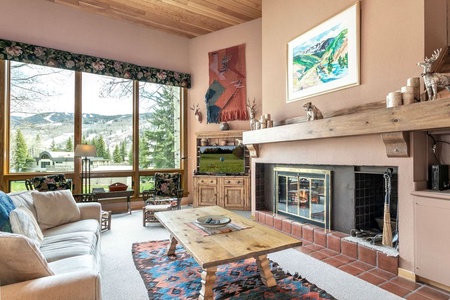 Cozy living room with large windows showing a mountain view, a fireplace, and a wooden coffee table. Sofa and armchairs are arranged around a patterned carpet. A TV is on the wooden cabinet.