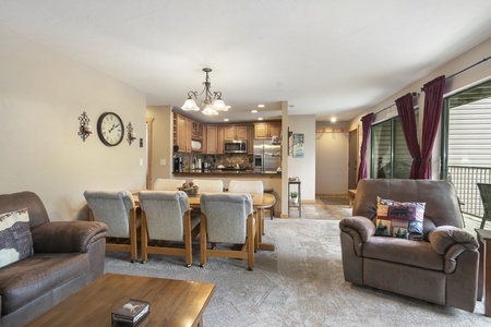 A cozy living and dining area with a central chandelier, a brown leather recliner, beige sofas, a wooden dining table with chairs, and a kitchen in the background. Maroon curtains adorn the windows.