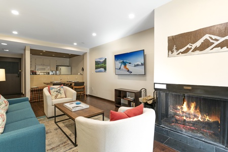 A cozy living room with a fireplace, two white armchairs, a blue sofa, a coffee table, and wall-mounted TV displaying a skiing scene. A kitchen area is visible in the background.