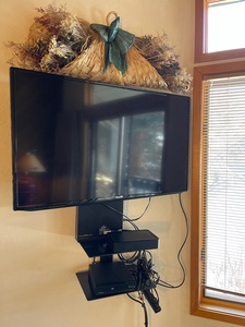 Wall-mounted flat-screen TV with attached shelves holding electronic devices and cables, underneath a decorative wicker basket with dried flowers and a ribbon. Wooden-framed window with blinds on the right.