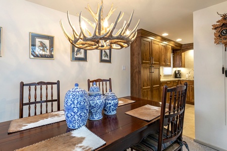 A dining room features a wooden table with blue ceramic jars, cowhide placemats, and an antler chandelier. Wooden chairs surround the table. The background shows framed artwork and the entrance to a kitchen.