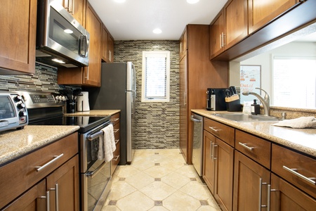 A modern kitchen with stainless steel appliances, wooden cabinets, and a tiled backsplash. A window is at the end of the room.