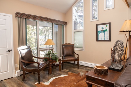 A small living room with two brown leather chairs, a wooden table, a cowhide rug, a lamp, a painting on the wall, and large windows with a nature view.