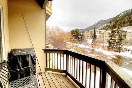 A small balcony with a grill, chair, and ski pole overlooks a snowy landscape with buildings and a mountain in the background.