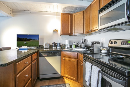 A modern kitchen with wooden cabinets, granite countertops, a stove, a microwave, and a dishwasher. A television displaying a golf course is placed on the counter.