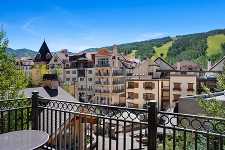 A scenic view of a European-style village with multiple buildings, balconies, and a clock tower against a backdrop of green hills and a clear blue sky.