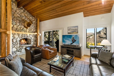 A cozy living room with a stone fireplace, leather seating, a large TV, and a wooden ceiling with beams. A window provides a view of a mountainous landscape.