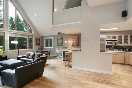 Open-plan living area with high ceilings, large windows, a modern kitchen, dining table, and black leather sofas. Colorful animal paintings decorate the walls, and light wooden flooring runs throughout.