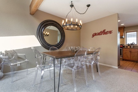 A dining area with a rectangular wooden table, clear plastic chairs, a round mirror, and a chandelier. The word "gather" is mounted on the wall. A kitchen is visible in the background.