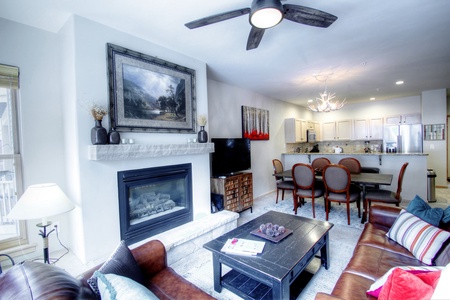 Modern living room and dining area featuring a fireplace, ceiling fan, sectional sofa, coffee table, and dining set. An open kitchen with bar seating is visible in the background.