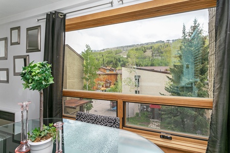 A large window with black curtains offers a view of a hillside with greenery. A dining table with a potted plant and glassware is in the foreground. Framed pictures hang on the adjacent wall.