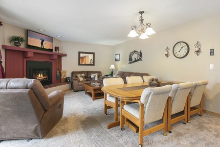 Living room with beige carpet, a wooden dining table and six chairs, a fireplace with a TV above it, a brown sofa set, and a framed clock on the wall.