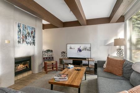 A cozy living room with a lit fireplace, a wall-mounted TV showing a snowy scene, and a wooden coffee table. The room has a large painting, a window with a view, and a bookshelf.