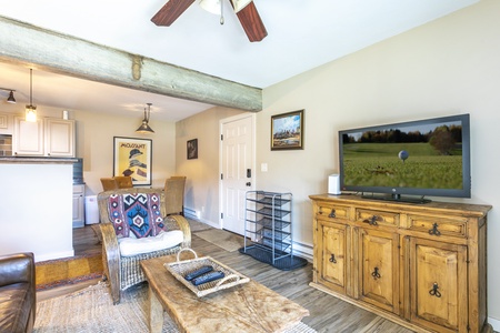 Living room with wooden furniture, a TV displaying a golf scene, a brown couch, a patterned blanket, and a view into a kitchen area. Ceiling fan and framed artwork are visible on the walls.