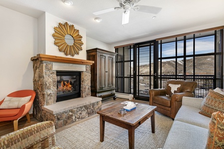 Cozy living room with a stone fireplace, brown leather armchair, beige couch, rustic wooden coffee table, and large windows with blinds showcasing a mountainous view.