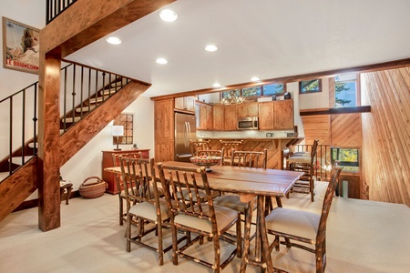 A rustic dining area with a wooden table and chairs, adjacent to a kitchen with wooden cabinets and stainless steel appliances. A staircase leads to an upper level. Natural light enters through high windows.