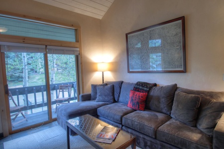 A living room with a gray sectional sofa, a red accent pillow, a coffee table with magazines, a floor lamp, a framed picture on the wall, and glass doors leading to a balcony with outdoor chairs.