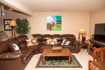 A cozy living room with a brown leather sectional sofa, a wooden coffee table, two table lamps, a TV on a stand, a potted plant, and wall art. The room has beige walls and a green rug.