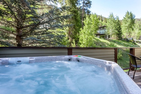 Hot tub on a wooden deck overlooking a lush, forested hillside with trees and a house in the background.