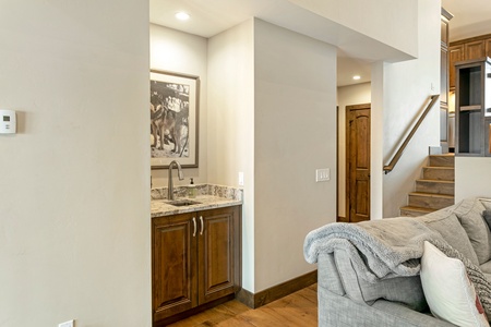 A small home bar area with a sink and two wooden cabinets is adjacent to a gray couch with blankets and a staircase leading upstairs. A framed picture hangs above the sink.