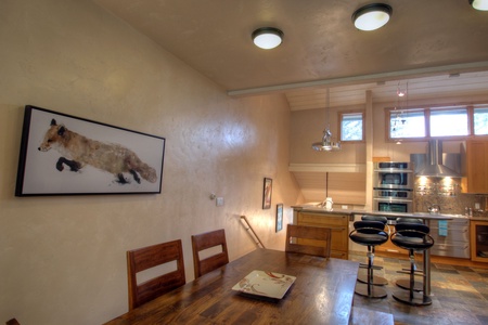 A modern kitchen and dining area featuring wooden furniture, a painting of a fox on the wall, and contemporary kitchen appliances under natural and artificial lighting.