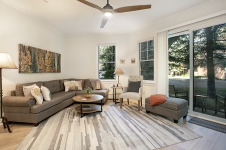 A cozy living room with a corner sofa, a round coffee table, an armchair, a standing lamp, and a ceiling fan. The room has large windows and a sliding glass door with views of outdoor greenery.