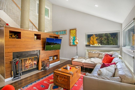 A cozy living room with a lit fireplace, modern decor, and a corner sofa with red and white cushions. A flat-screen TV is mounted above the fireplace, and a large window shows a forest view.