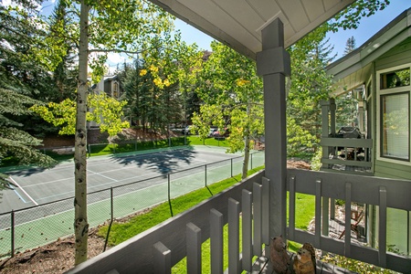 View from a balcony overlooking a green fenced tennis court surrounded by trees and houses under a clear sky.