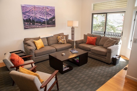 A cozy living room with two gray sofas, two armchairs, a wooden coffee table, a standing lamp, and a mountain landscape painting on the wall. The window offers a view of trees outside.