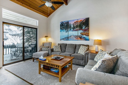 A cozy living room with gray sofas, a wooden coffee table, a ceiling fan, and a large painting on the wall. A sliding glass door opens to a snowy outdoor area with a table and chairs.