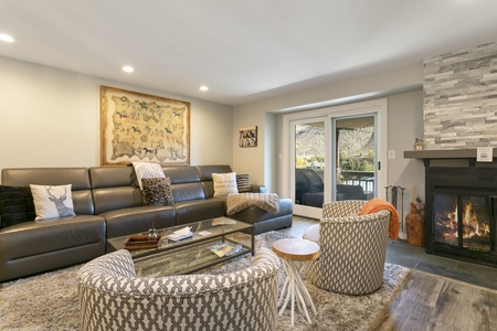 A cozy living room with a long, dark leather sofa, two patterned chairs, a wooden coffee table, a fireplace, and a wall map art piece. There are glass doors opening to an outdoor area.