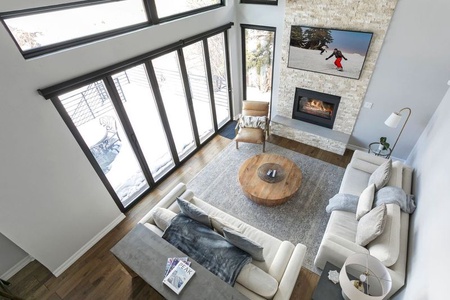 A modern living room with a gray sofa, wooden coffee table, and a fireplace. A person snowboarding is shown on the wall-mounted TV above the fireplace. Large windows showcase a snowy outdoor view.