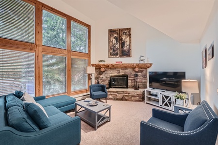 A living room features a stone fireplace, large windows with blinds, blue furniture, a TV on a white stand, and a coffee table. The decor includes wall art, lamps, and small decorative items on the mantle.