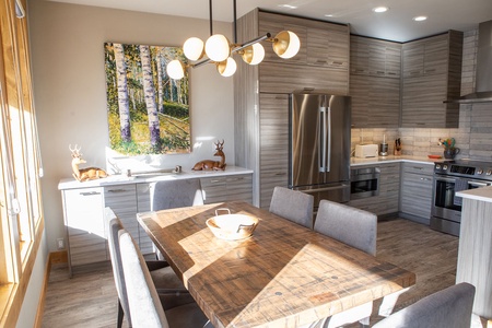 Modern kitchen with wooden dining table, gray upholstered chairs, stainless steel appliances, light wood cabinetry, and a painting of birch trees on the wall. Bright natural light from a window.