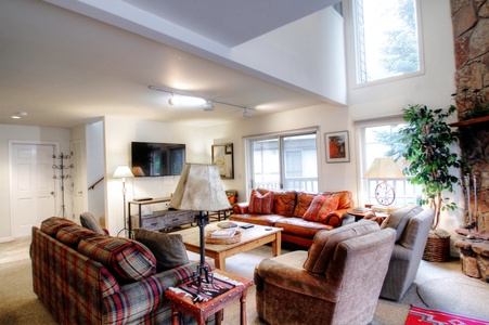 A cozy living room with various seating options, including a red couch, plaid sofa, and beige armchairs. The room has a fireplace, large windows, a wall-mounted TV, a small wooden table, and lamps.