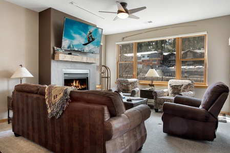 A living room with a fireplace, wall-mounted TV, and a large window. There are two brown sofas and two patterned armchairs arranged around a coffee table. A ceiling fan is on the white ceiling.