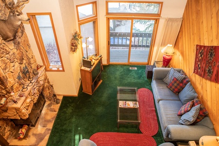 A cozy living room with a stone fireplace, green carpet, and a blue sofa with plaid cushions. A mounted deer head is above the fireplace, and sliding glass doors lead to an outdoor balcony.