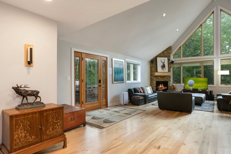 A spacious living room with wooden flooring, a stone fireplace, large windows, and a glass front door. The room features modern furniture and nature-themed decorations, including a sculpture and paintings.