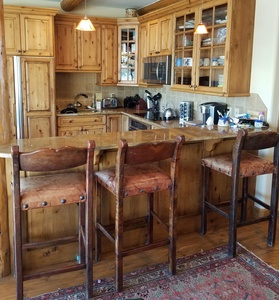 A rustic kitchen with wooden cabinets and a countertop bar featuring three wooden stools. The kitchen has hanging lights, a stove, microwave, a coffee maker, and other kitchen appliances.