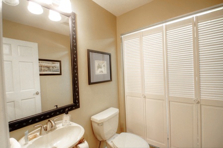 A bathroom with a toilet, sink, and mirror. Wall art is visible next to a window with closed blinds. A door is partly open on the left.